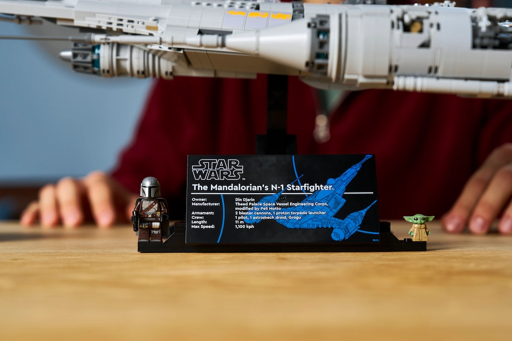 Display plaque for Star Wars: The Mandalorian's N-1 Starfighter with a Mandalorian minifigure and Grogu on a wooden table, hands blurred in the background.