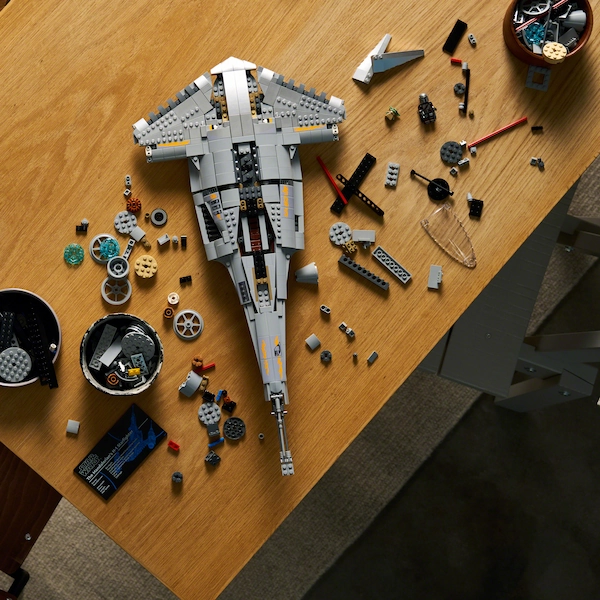 A gray LEGO spaceship under construction on a wooden table, with scattered bricks, gears, and small parts nearby in bowls and on the surface.
