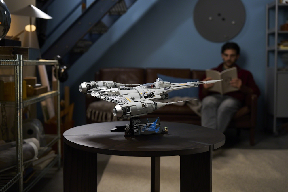 LEGO Mandalorians N-1 Starfighter model on a round table in a cozy room, with a man reading a book in the background on a sofa.